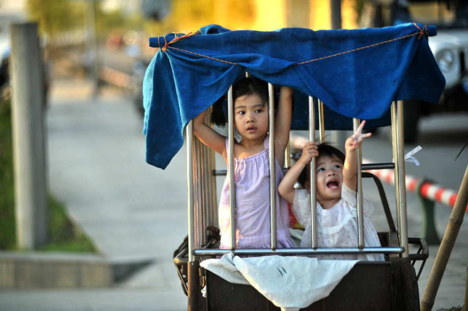 Two girls who look like 6 and 3 years old respectively are locked in a cage-like tricycle on roadside, while their grandmother is doing laundry by the river in S China’s Guangxi June 18, 2013.(Photo/ youth.cn)